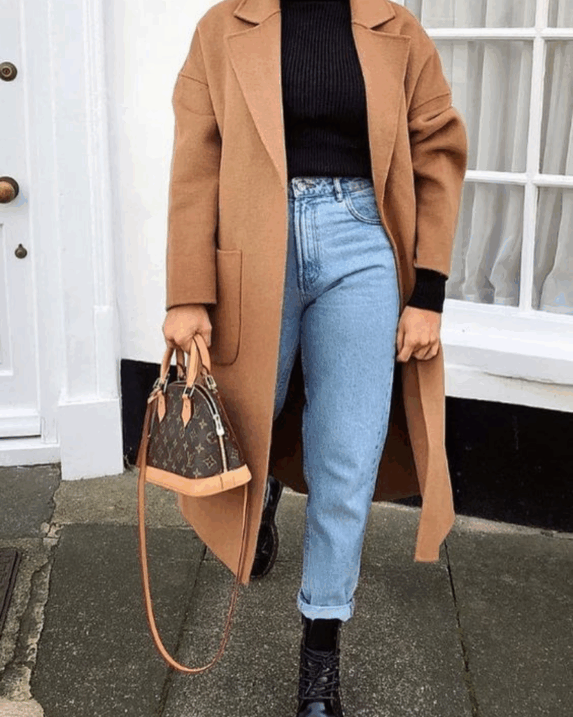 Woman wearing a camel coat, high-waisted light-wash jeans, black ribbed top, and black lace-up boots while holding a Louis Vuitton handbag.
