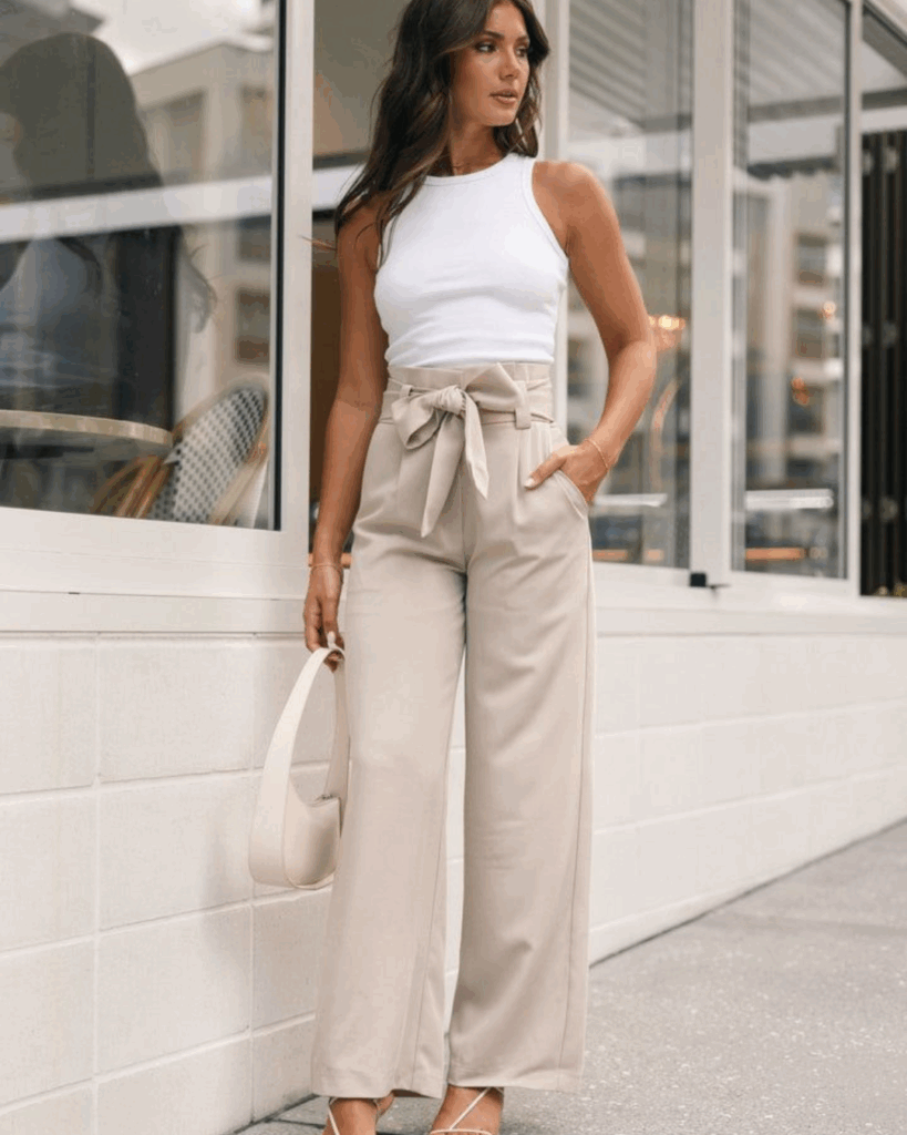 Woman in a white tank top and high-waisted beige tie-front trousers, standing outdoors with a modern and casual chic vibe.