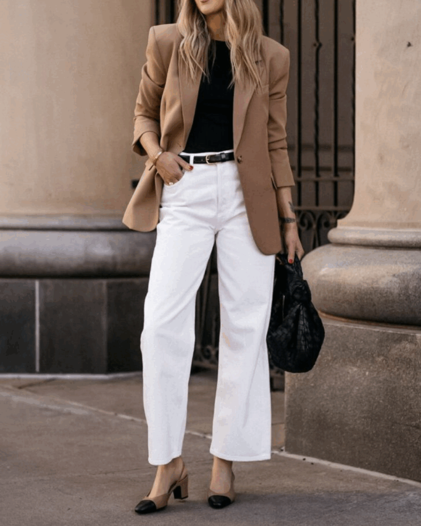 A woman styles a camel blazer over a black top with high-waisted white jeans and two-tone shoes, carrying a black handbag.