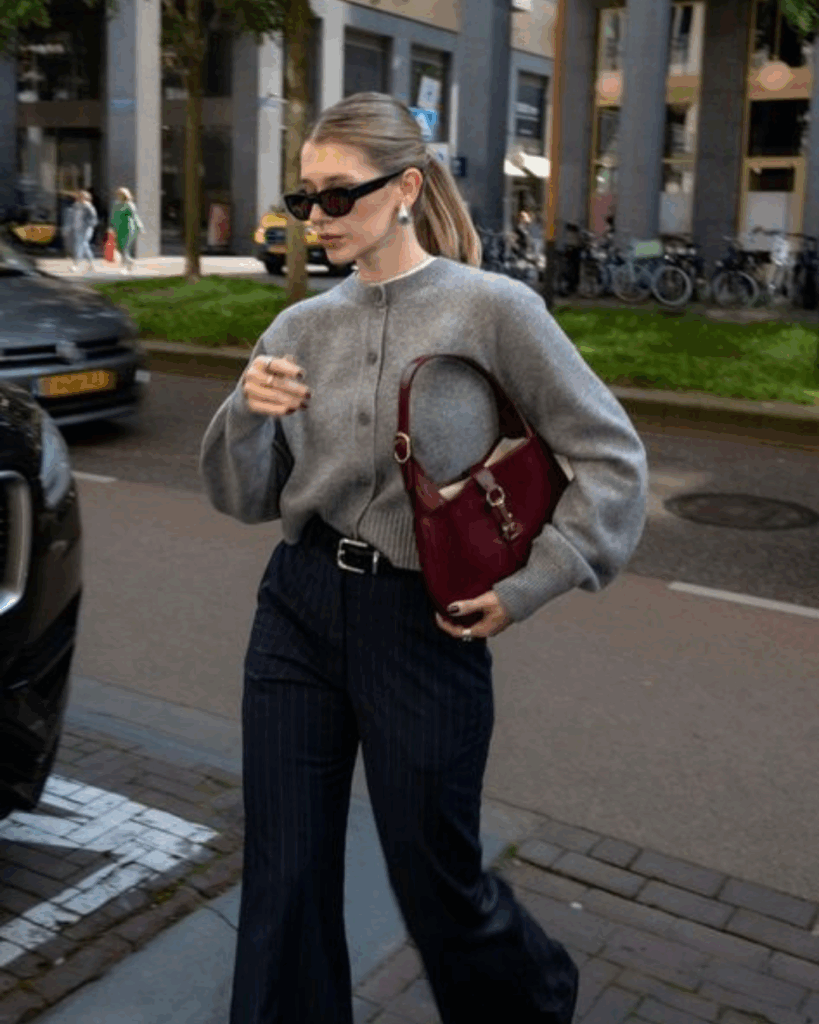 Woman wearing a grey cardigan tucked into navy striped trousers, accessorized with sunglasses and a burgundy handbag.
