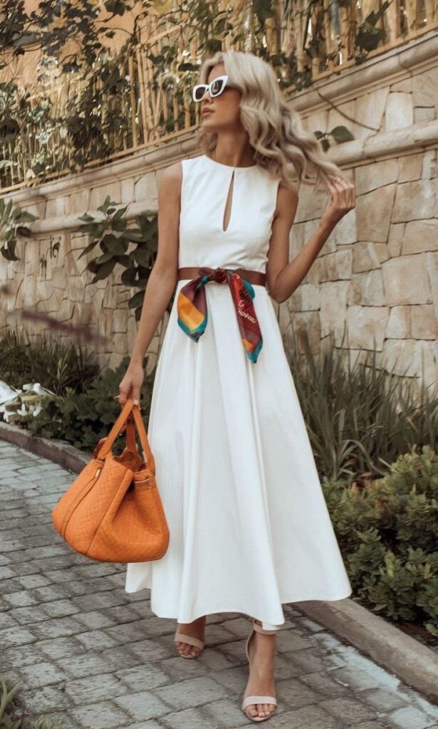 Woman in a sleeveless white midi dress styled with a colorful scarf belt and orange handbag.