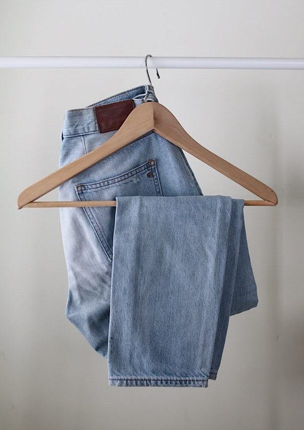 A pair of light wash blue jeans folded over a wooden hanger, hanging on a white clothing rack against a minimalist background.
