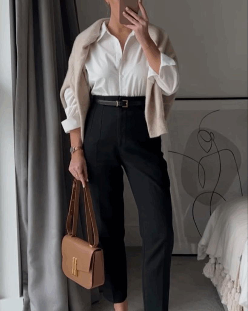 Woman wearing a smart casual outfit with a white button-down shirt, black tailored trousers, beige sweater draped over shoulders, and tan crossbody bag.
