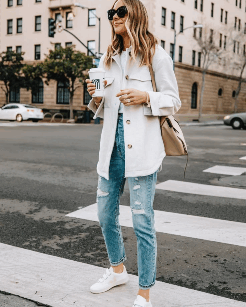Stylish woman crossing a city street wearing a white button-up shacket, distressed jeans, white sneakers, and sunglasses while holding a coffee cup.