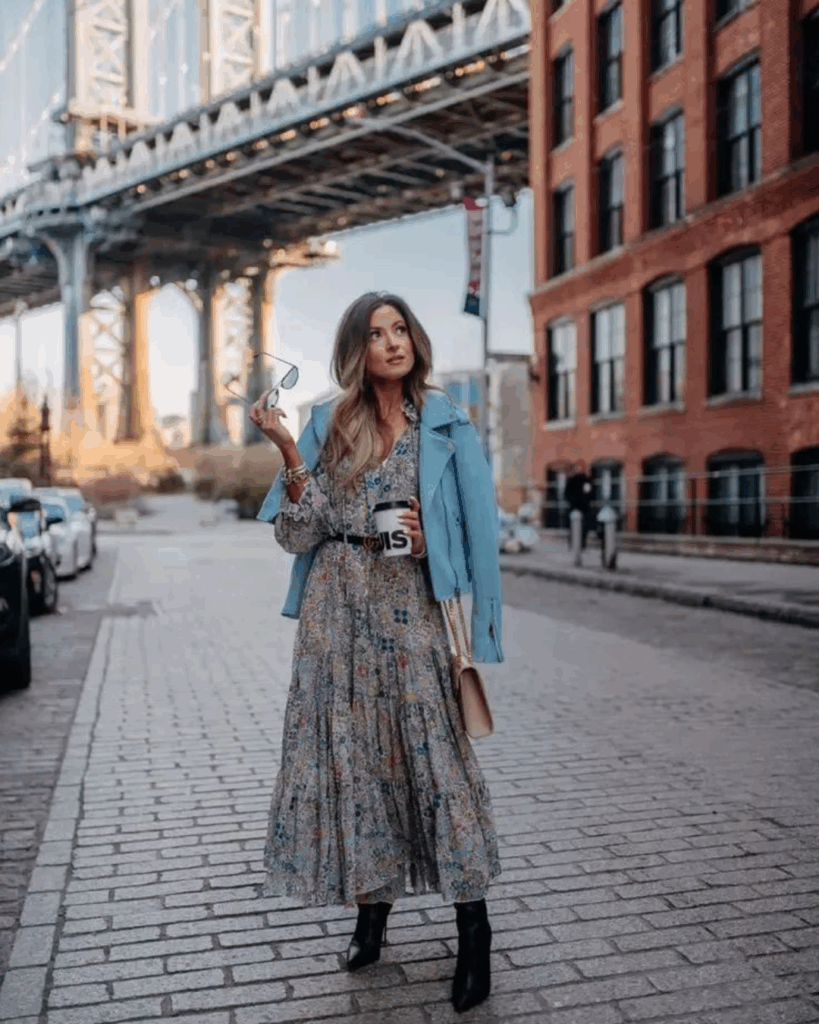 A stylish woman stands on a cobblestone street under a bridge, wearing a floral maxi dress, a light blue jacket draped over her shoulders, black ankle boots, and holding sunglasses and a coffee cup.