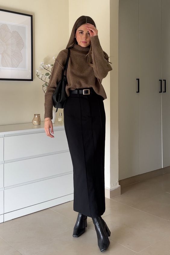 Woman wearing a brown turtleneck sweater tucked into a black midi skirt with belt, black boots, and a shoulder bag indoors