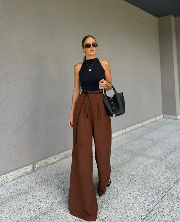 Stylish woman wearing a sleeveless black top with high-waisted wide-leg brown trousers, accessorised with sunglasses, gold jewellery, and a black handbag.