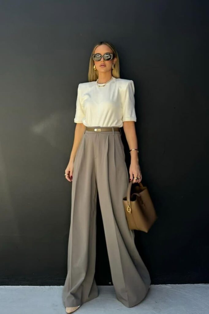 A woman stands against a black wall wearing a structured white blouse tucked into high-waisted taupe wide-leg trousers, accessorized with nude pointed-toe heels, oversized sunglasses, pearl jewelry, and a brown structured handbag.
