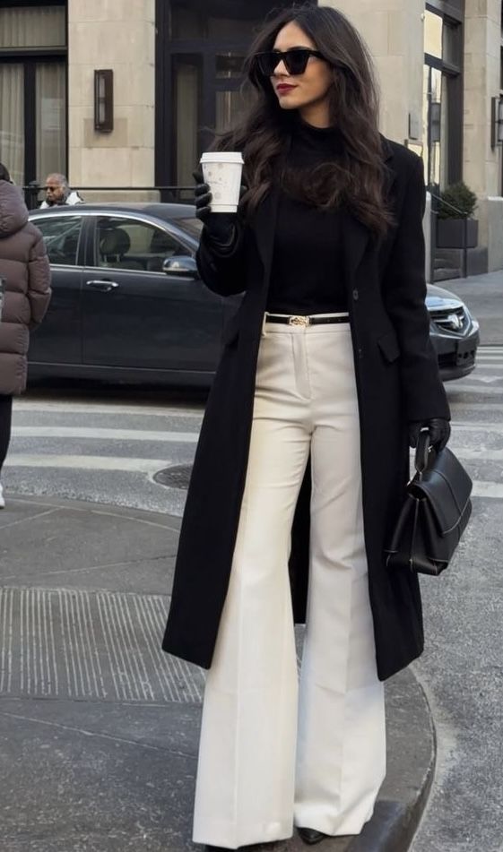 A woman crosses the street wearing a long black coat over a black turtleneck and high-waisted white wide-leg trousers, accessorized with pointed black heels, oversized sunglasses, black gloves, and a structured black handbag.