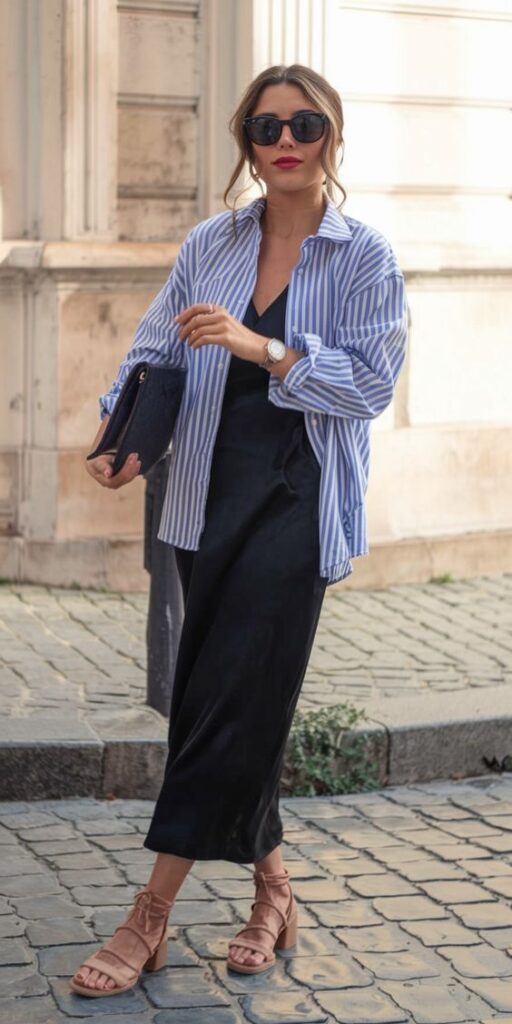Woman wearing a blue striped shirt over a black slip dress with tan block-heel sandals and a black clutch.