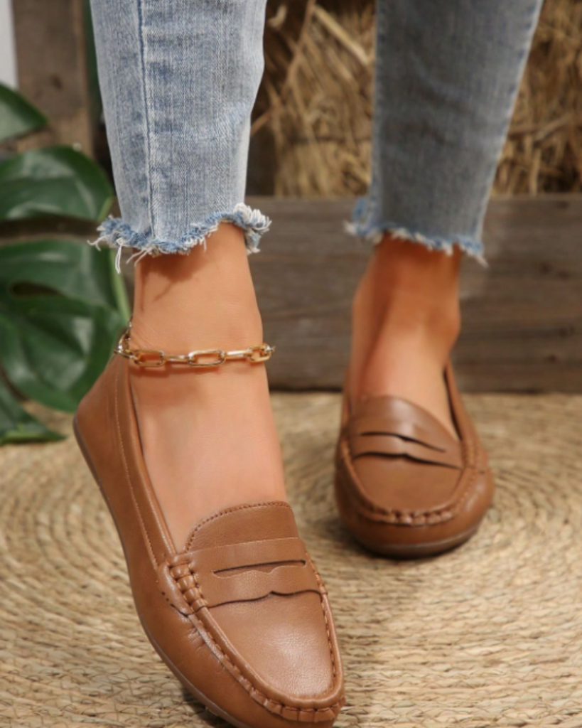 Tan leather loafers with gold chain detail styled with light wash cropped jeans on a woven rug.