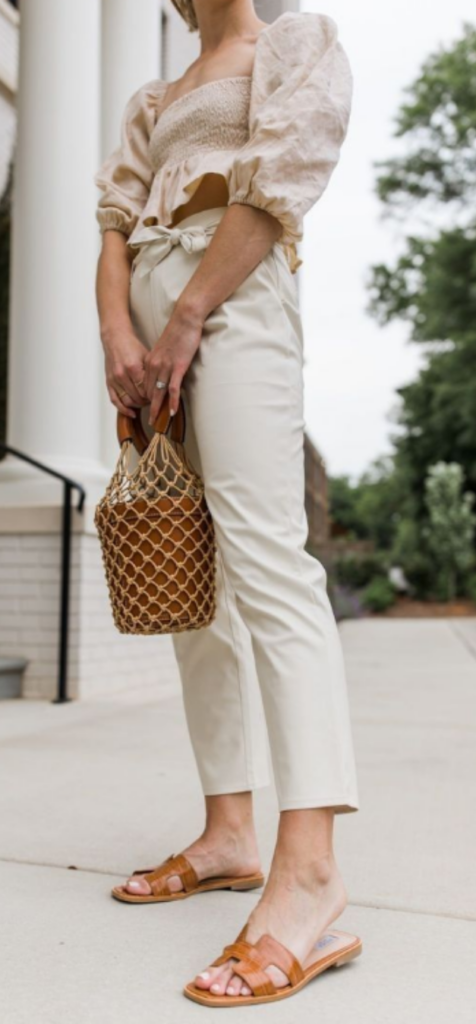 Woman wearing minimal tan strappy sandals with neutral tailored trousers and woven bag.